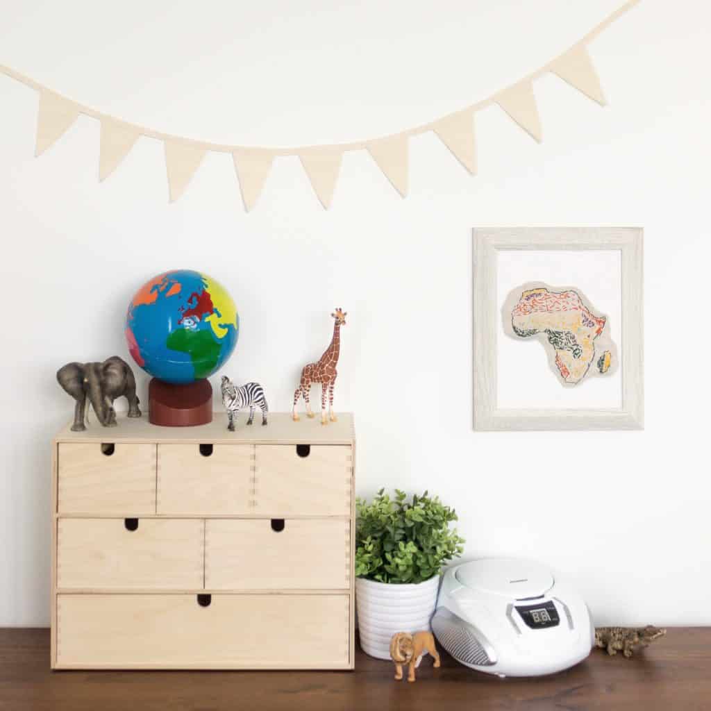 A continents globe sits on top of a Moppe storage unit from IKEA. An embroidered biome map sewn by a child is hung on the wall, and several Schleich animals are displayed beside it, inviting open-ended play and exploration for young children. 