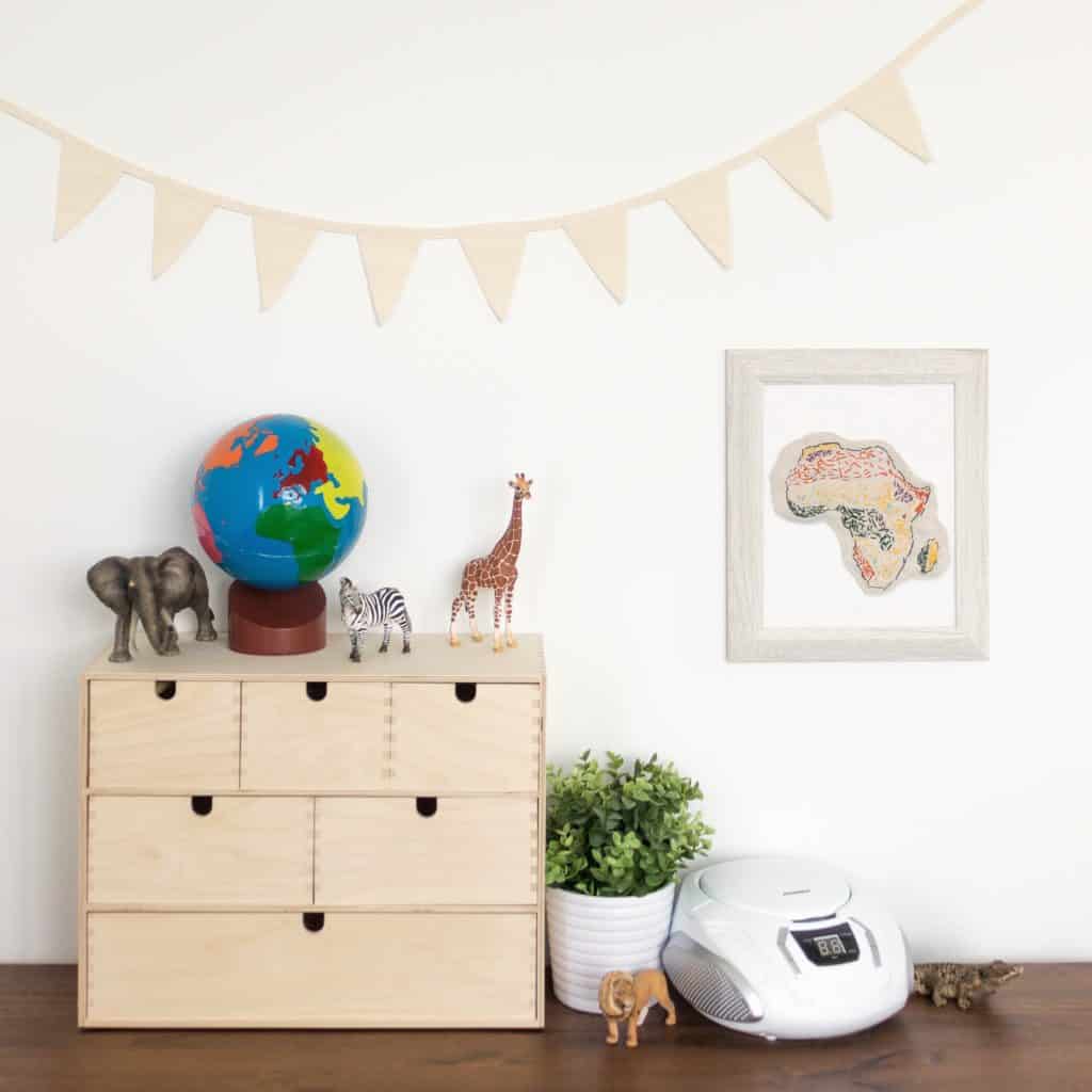 A continents globe sits on top of a Moppe storage unit from IKEA. An embroidered biome map sewn by a child is hung on the wall, and several Schleich animals are displayed beside it, inviting open-ended play and exploration for young children.